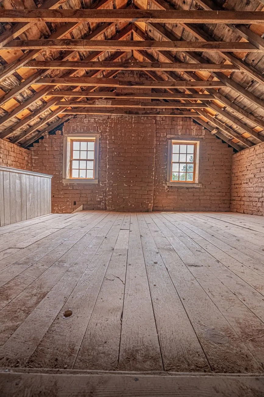 A converted loft with exposed brick gable wall, two small windows and a wide boarded floor under the roof timbers